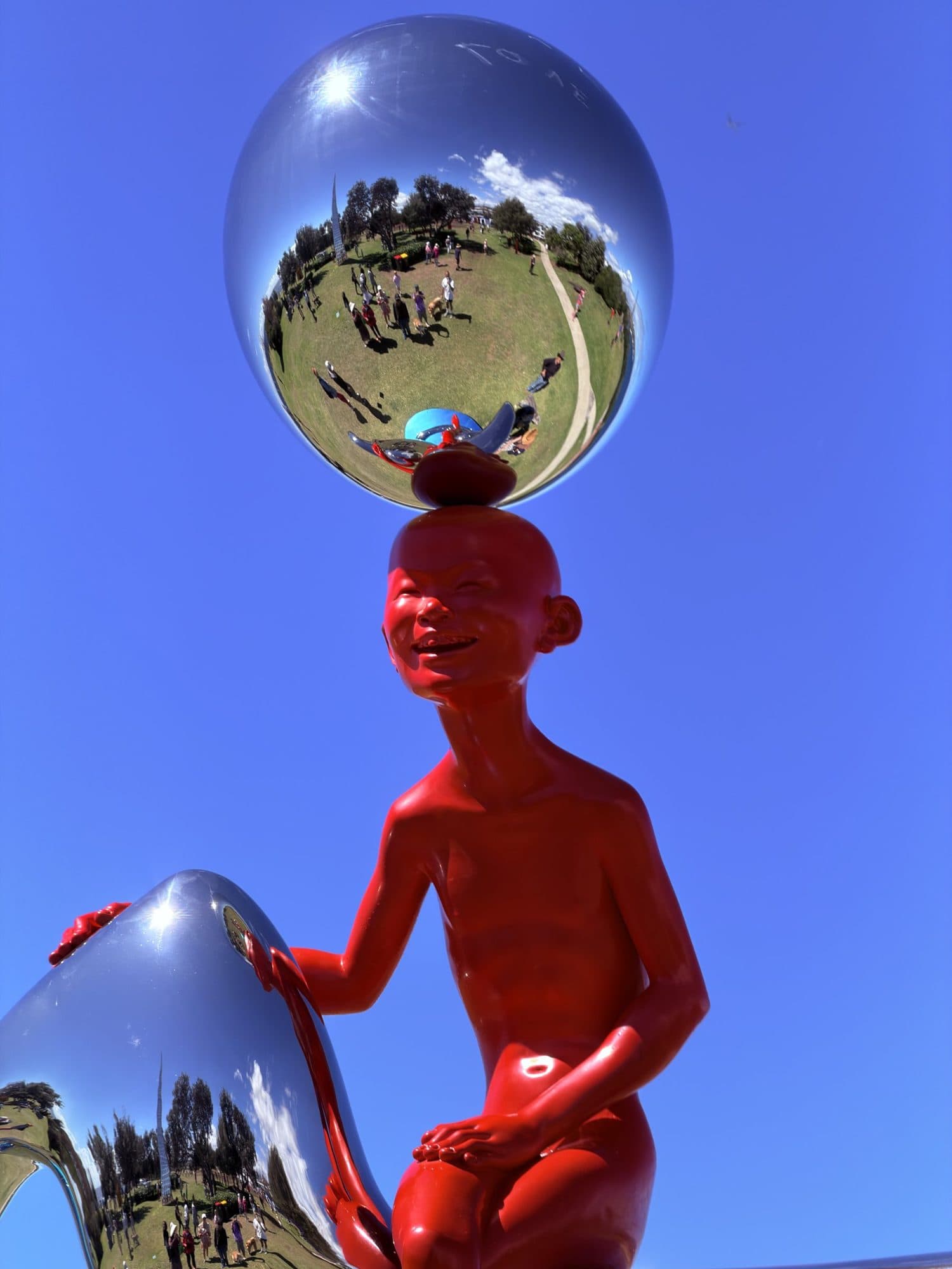 Chen Wenling's Cloud Watcher: a grinning red figure holding a mirrored sphere above his head, reflecting the crowd and lawn of Marks Park, against a blue sky