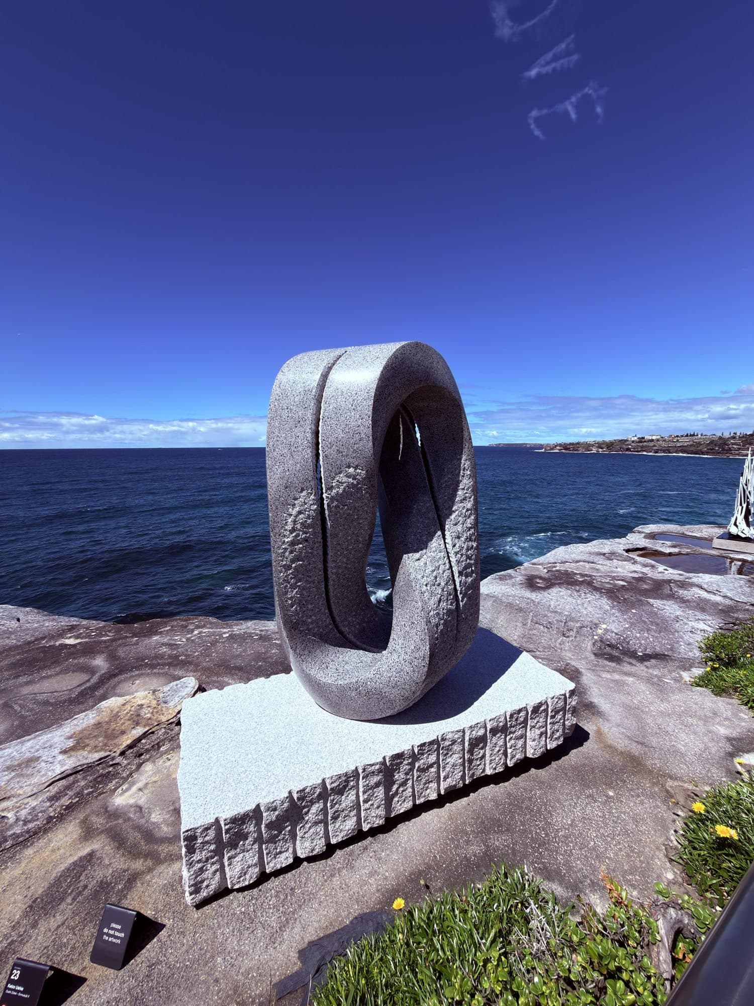 Keizo Ushio's grey granite twisted loop on a white granite base, on a sandstone ledge above the ocean with the coastline behind