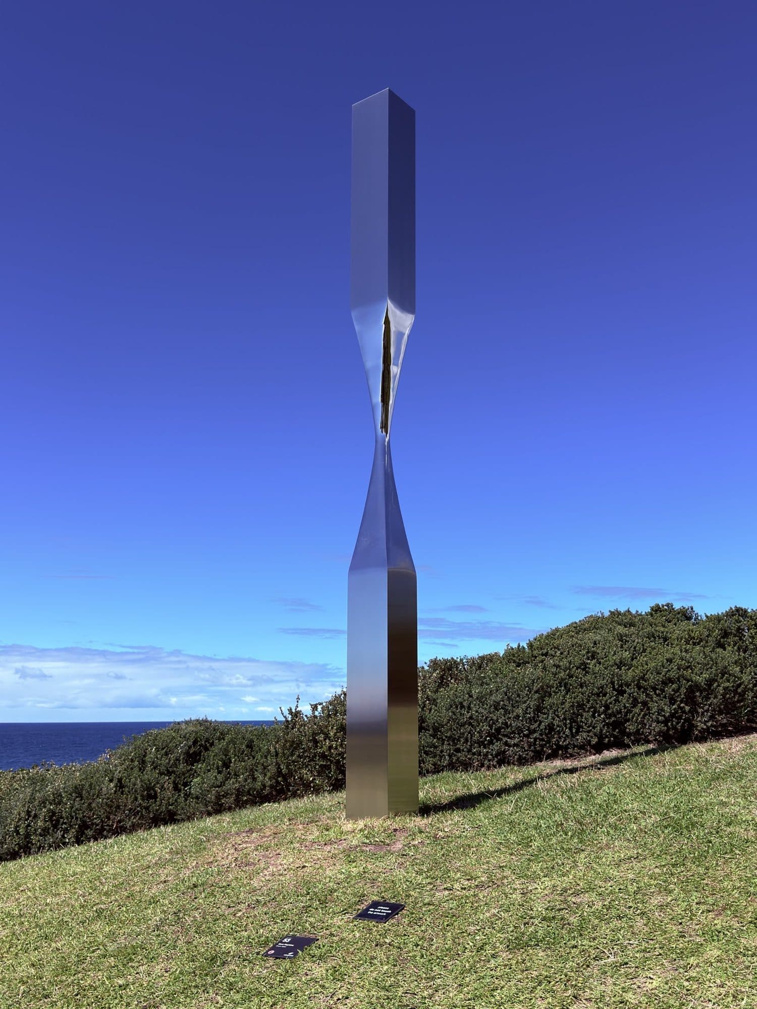 Second view of Sam Hopkins' Drawn Apart: a mirrored steel column pinched in the middle, on a grassy slope above the sea