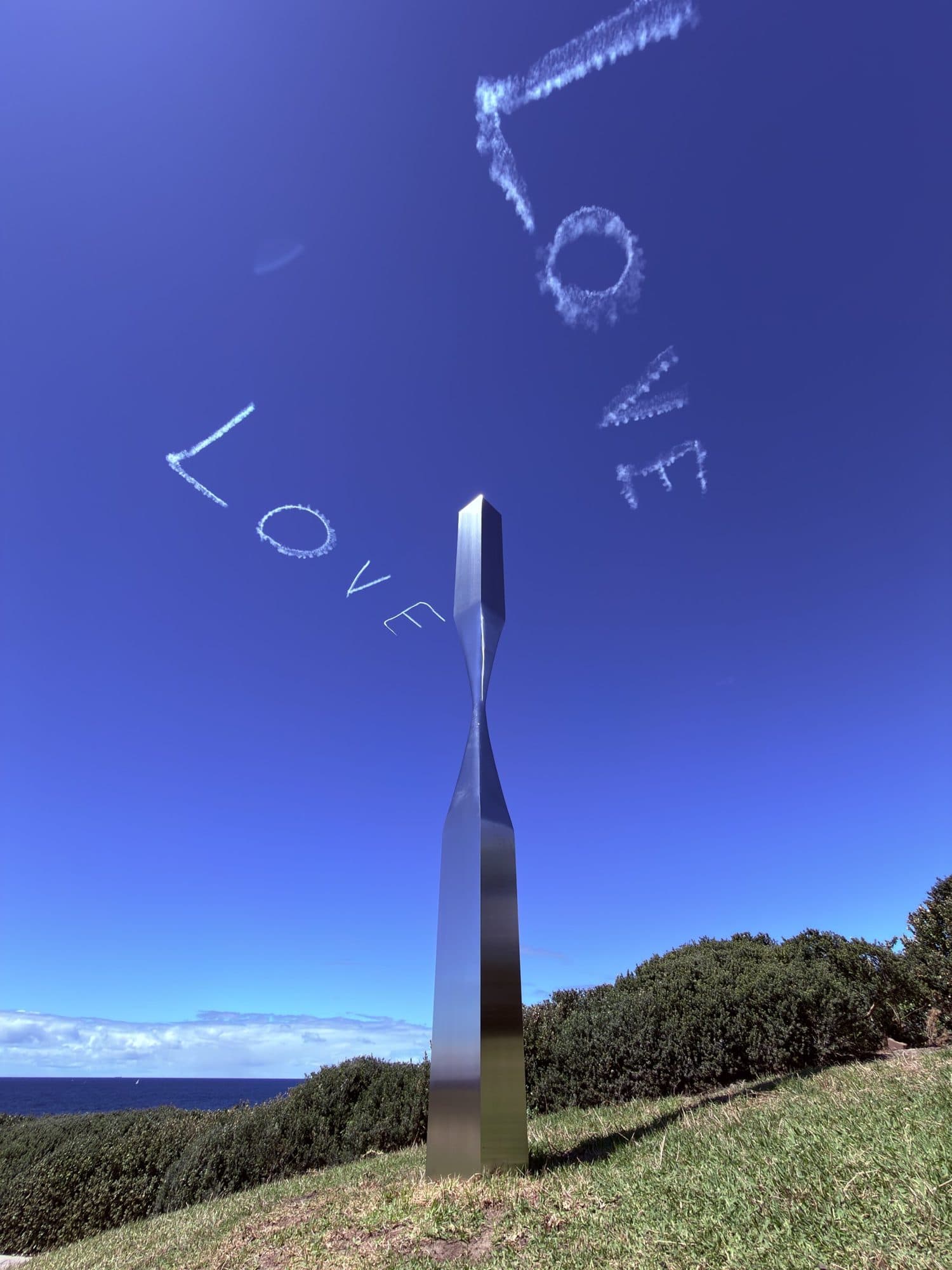 Sam Hopkins' Drawn Apart: a slender mirrored steel column pinched in the middle, with the word LOVE skywritten in the blue sky above it