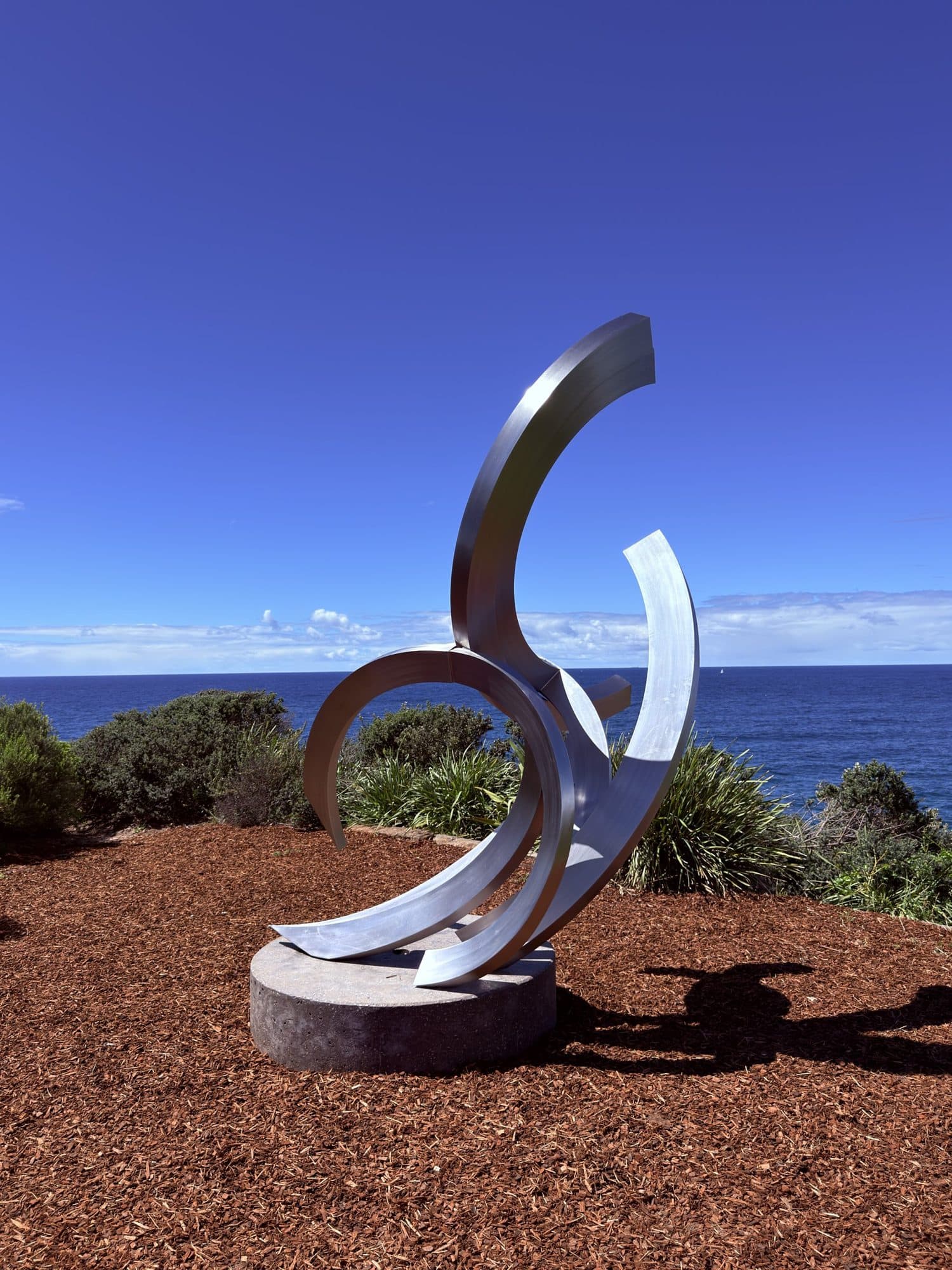 James Parrett's M-fiftyseven: sweeping polished steel arcs curling upward like a wave, on a bed of red mulch with the ocean behind
