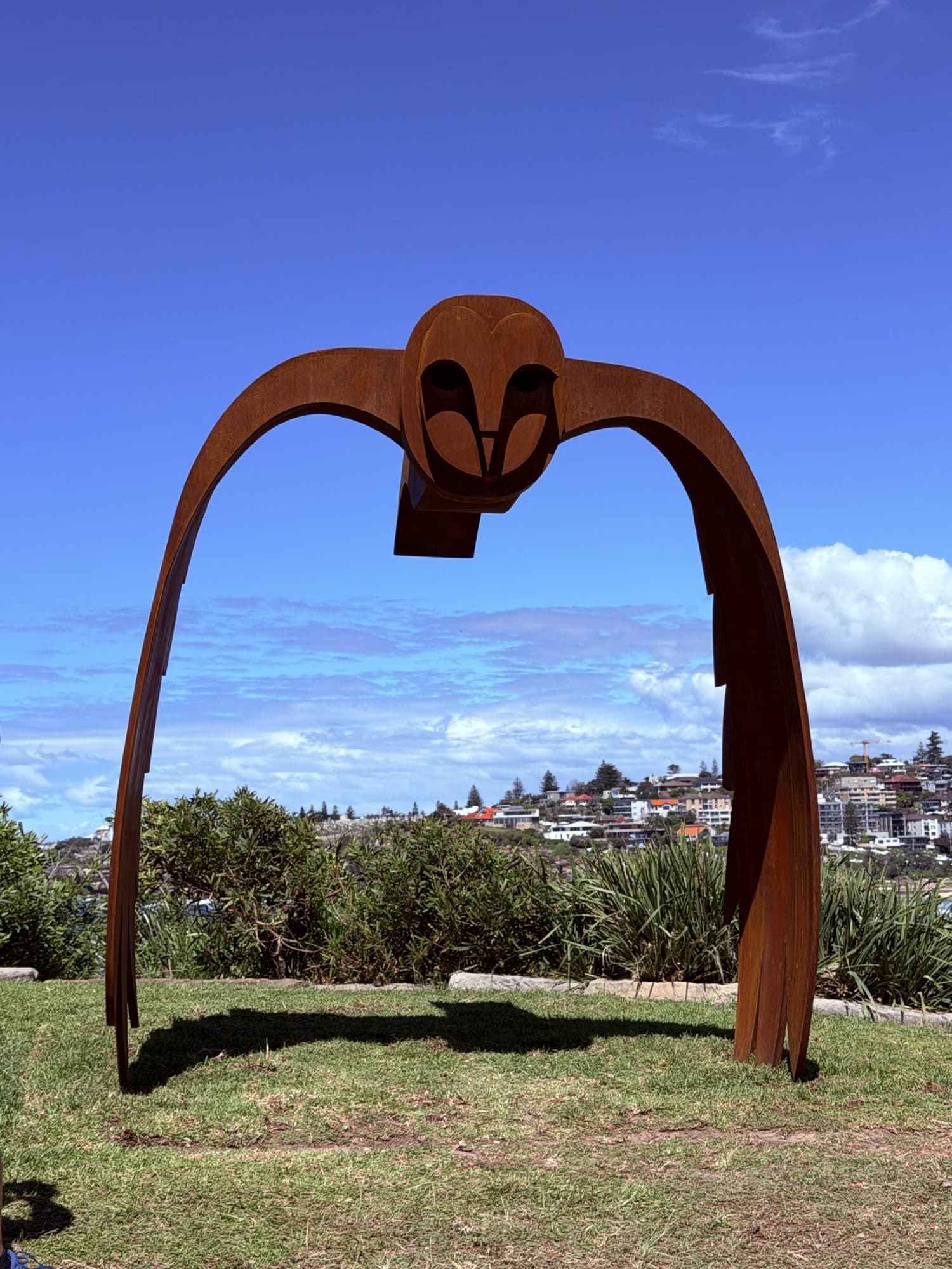 Jimmy Rix's Silent Crossing: a rust-coloured steel owl with long arched wings standing on the lawn, Bondi apartments behind