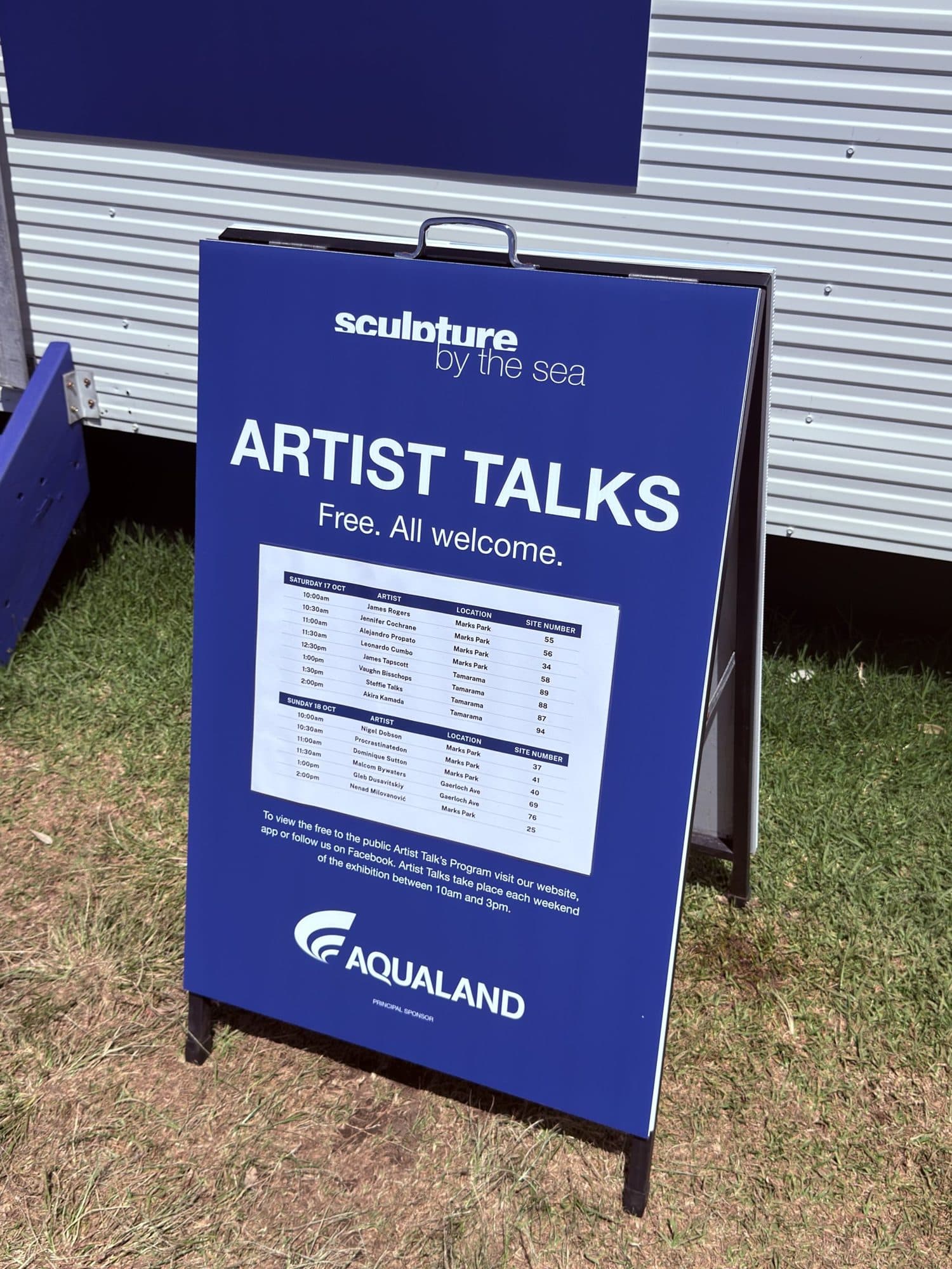 Blue Aqualand Artist Talks board on the coastal walk listing opening weekend times, artists, locations and site numbers